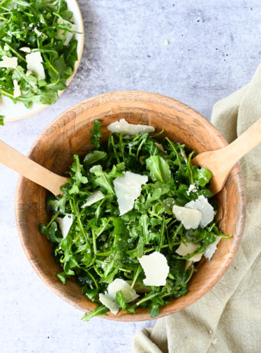 Utensils in a wooden bowl of arugula salad with lemon, olive oil, and Parmigiano-Reggiano.
