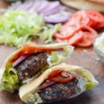 Greek-style lamb burgers on a wooden surface with burger toppings.