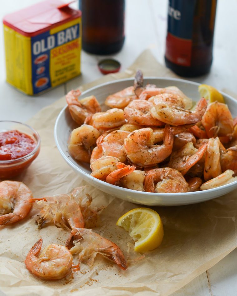 Bowl of peel-and-eat boiled shrimp with cocktail sauce in from of Old Bay Seasoning.