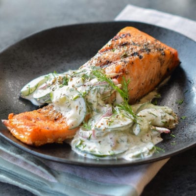 Grilled salmon topped with cucumber-dill salad on a black plate.