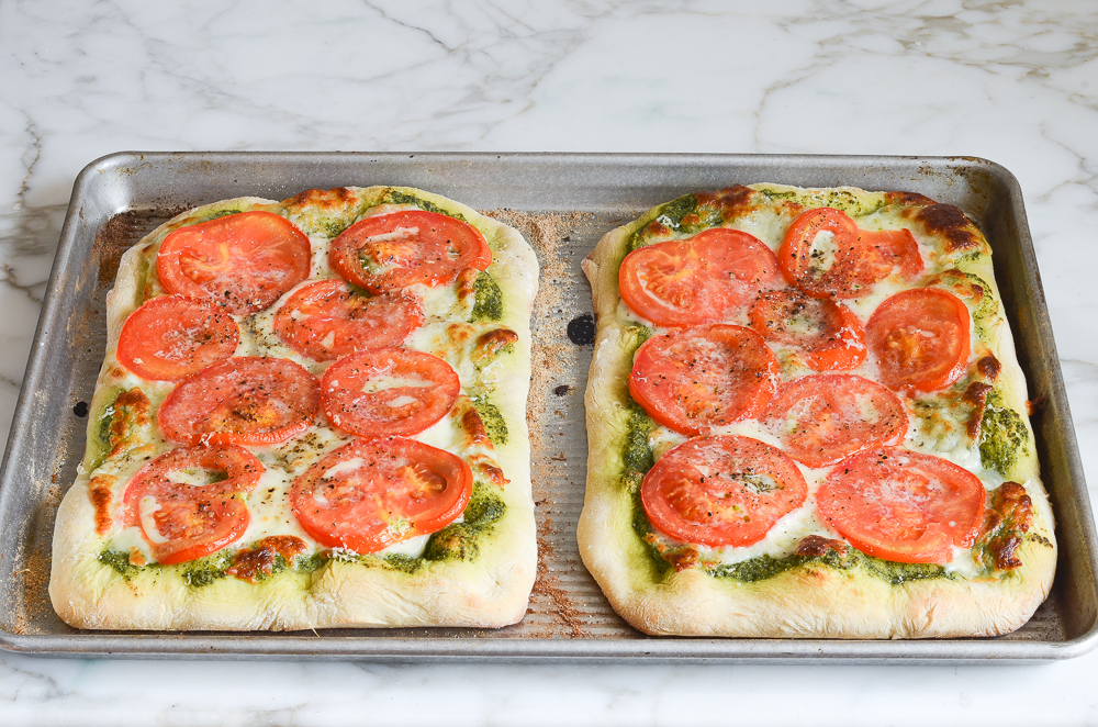 Two pesto pizzas with fresh tomatoes and mozzarella on a baking sheet.