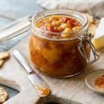 Jar of roasted tomato peach jam on cutting board with wedges of cheese.