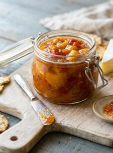 Jar of roasted tomato peach jam on cutting board with wedges of cheese.