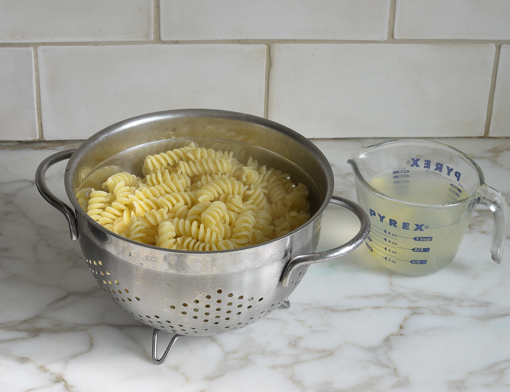 Colander of pasta next to a measuring cup of pasta water.