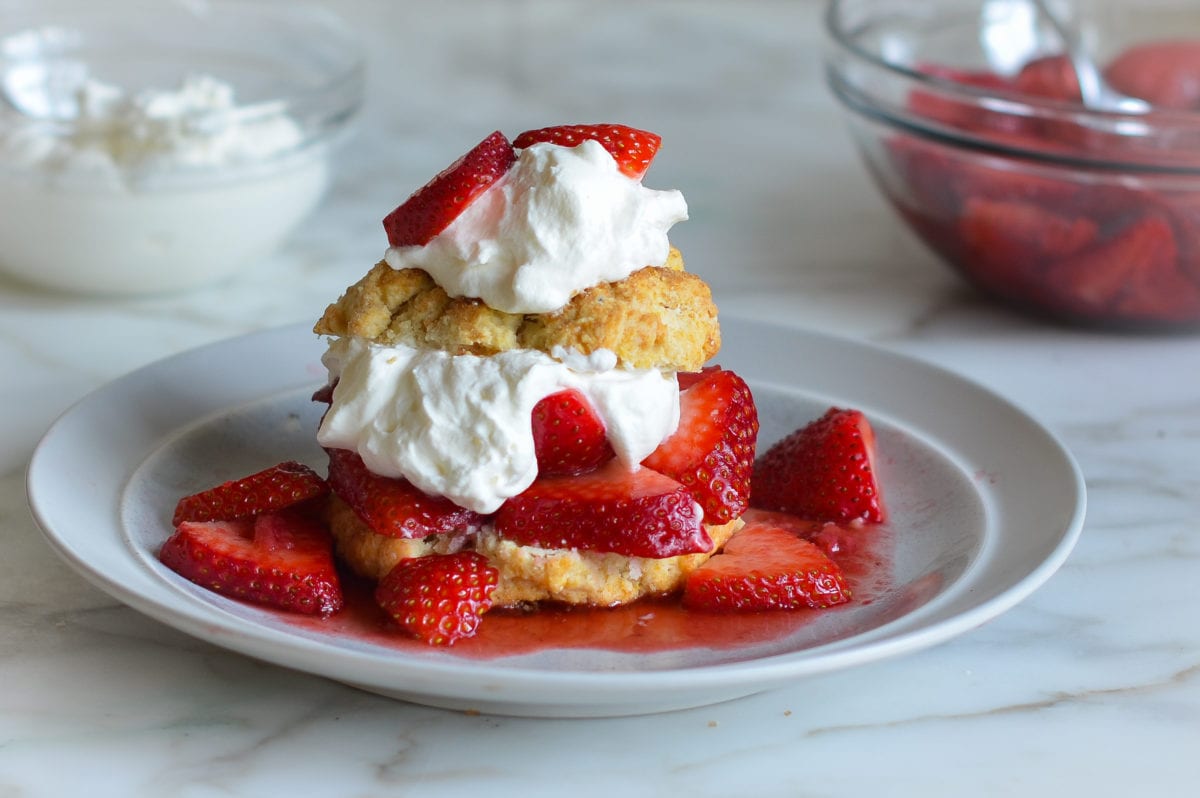 strawberry shortcake on plate with bowls of strawberries and whipped cream