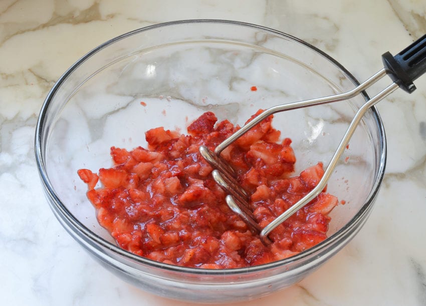 Potato masher in a bowl of pureed strawberries.