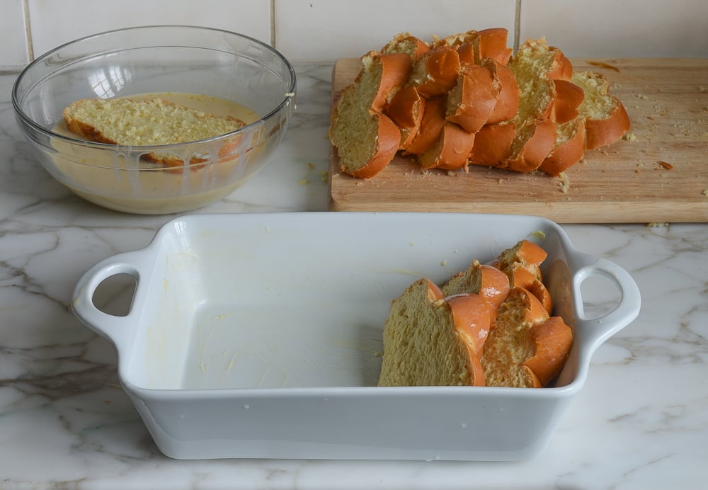 Slice of challah in a bowl of custard next to a baking dish.
