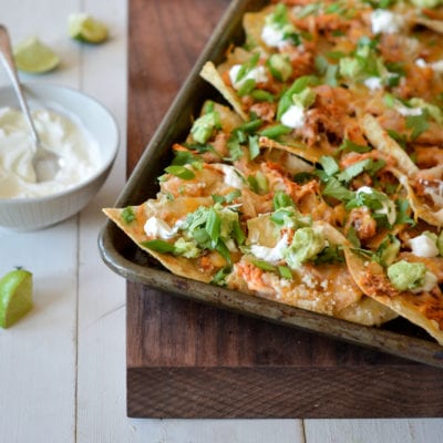 Sheet pan chicken nachos on a wooden board with a bowl of sauce and lime wedges to the side.