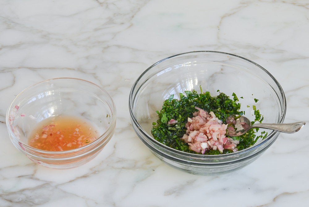 Small bowl of vinegar next to a bowl of herbs and shallots.