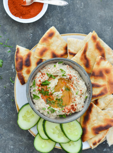 Bowl of baba ganoush on a plate with cucumbers and bread.