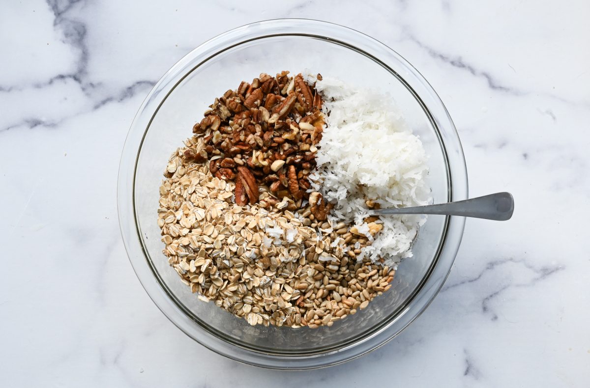 oats, nuts, seeds, and shredded coconut added to wet ingredients in bowl