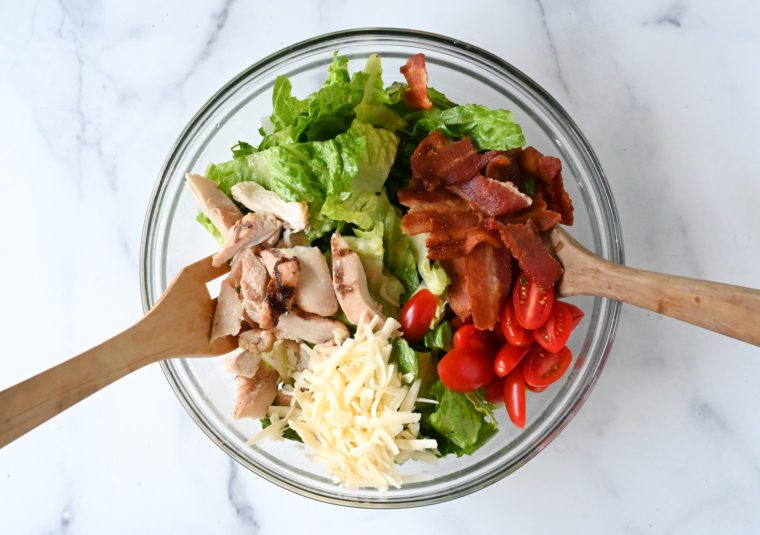 adding bacon, chicken, tomatoes and cheese to dressed greens