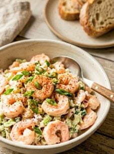 Shrimp salad in white bowl on wooden table with toast in background.