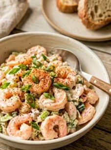 Shrimp salad in white bowl on wooden table with toast in background.