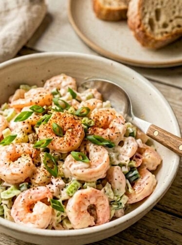 Shrimp salad in white bowl on wooden table with toast in background.