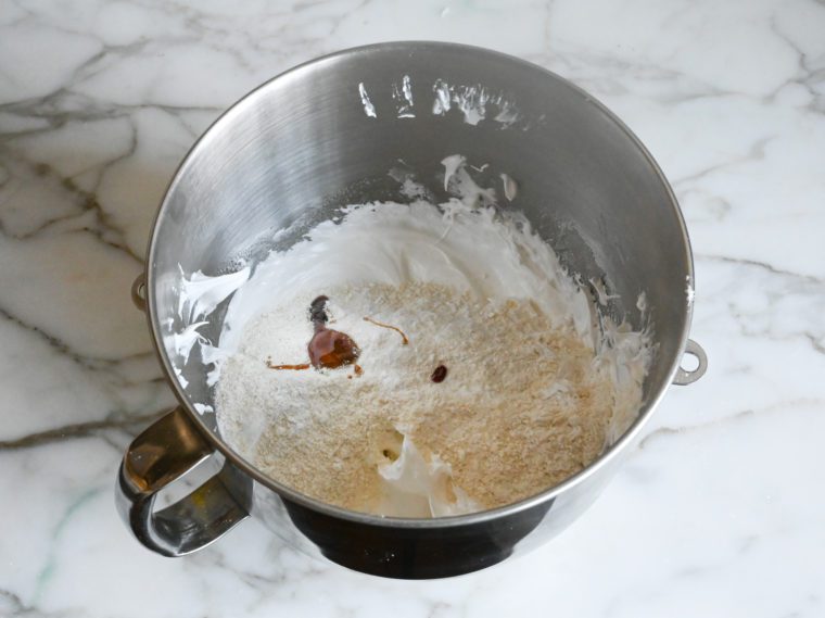 adding the almond flour and confectioners sugar mixture, food coloring, and vanilla paste to the meringue in the mixing bowl