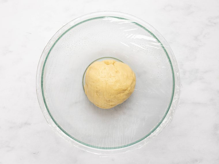 Ball of dinner roll dough in a bowl covered with plastic wrap.