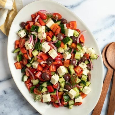 A large platter of Greek salad on a table with a wooden spoon to the side.