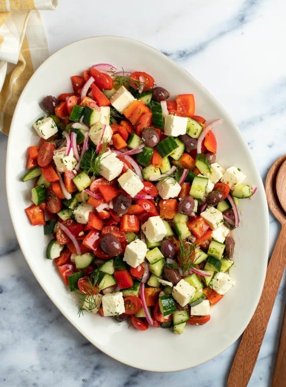A large platter of Greek salad on a table with a wooden spoon to the side.