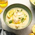 Avgolemono soup in bowl with gray striped dish towel, olive oil, and lemon wedges.