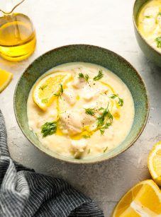 Avgolemono soup in bowl with gray striped dish towel, olive oil, and lemon wedges.