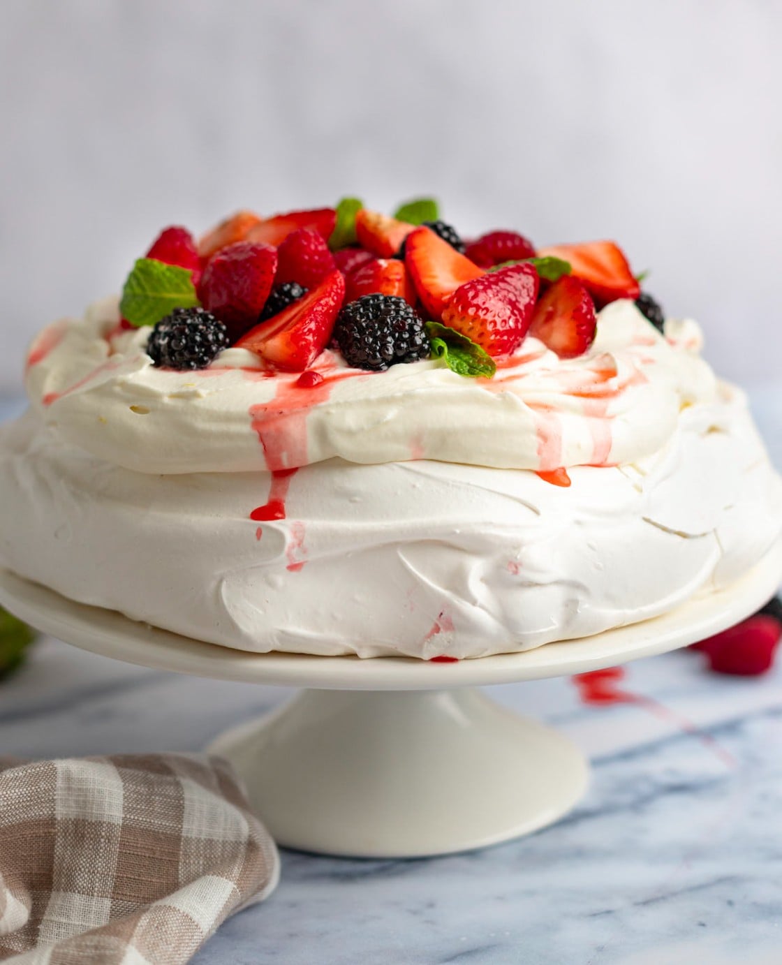 Pavlova topped with macerated berries on a cake stand with a linen napkin to the side.