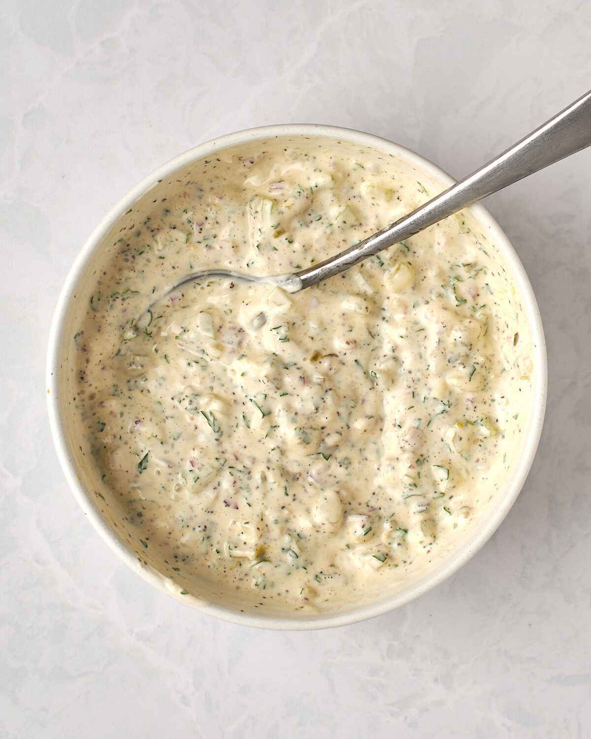 large white bowl with spoon combining celery, onion, relish, mayonnaise, dry mustard, celery seed, parsley, pepper, and salt