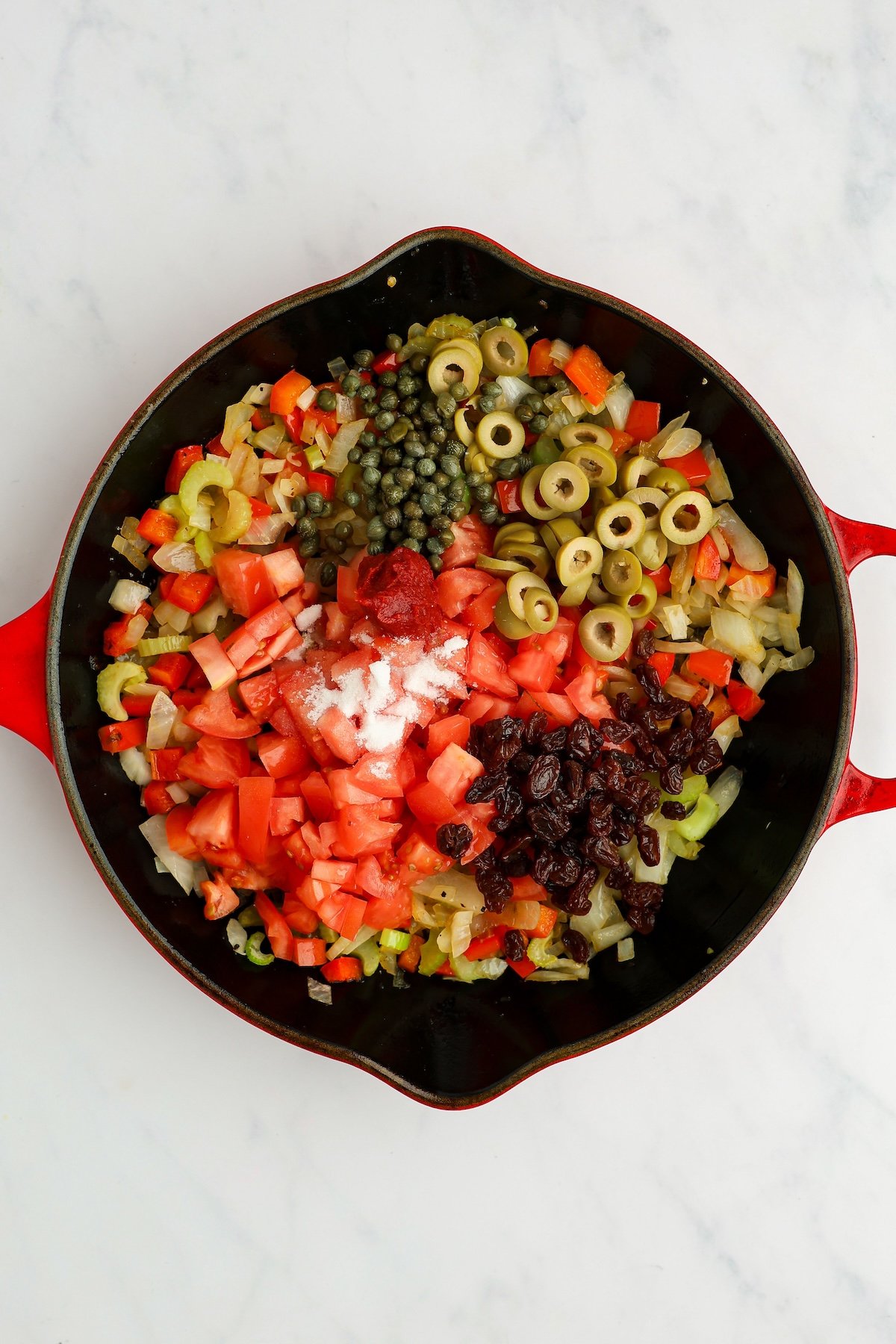 large skillet with tomatoes, tomato paste, olives, raisins, capers, and sugar added to celery, onion, and bell pepper