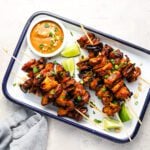 white serving platter with cooked chicken skewers and small bowl of peanut sauce