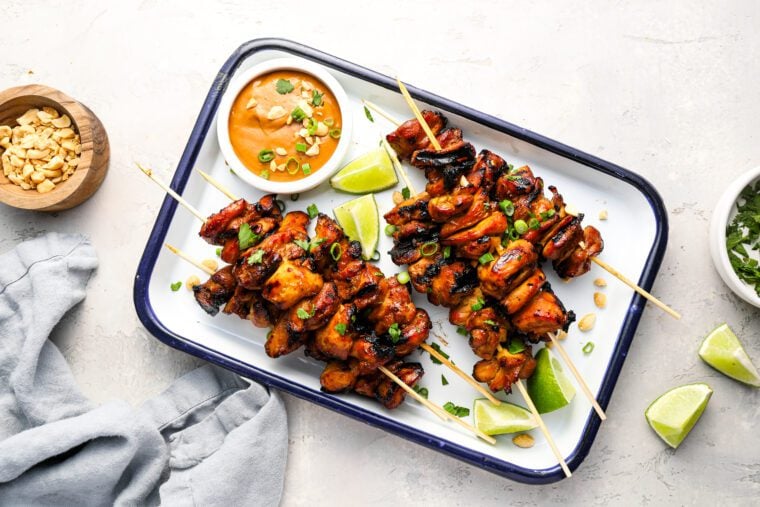 white serving platter with cooked chicken skewers and small bowl of peanut sauce