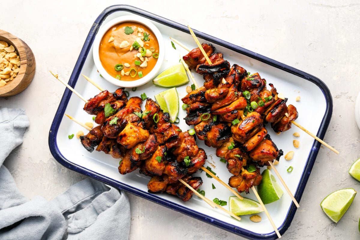 white serving platter with cooked chicken skewers and small bowl of peanut sauce