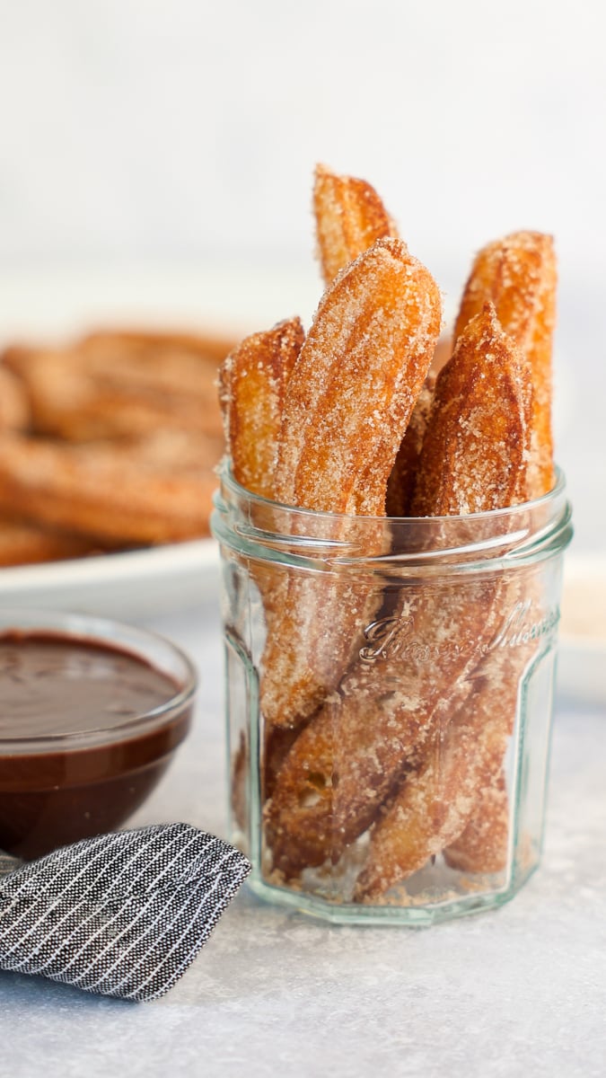churros in glass jar with chocolate sauce and platter of churros in background.