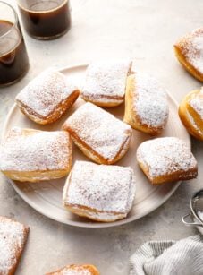 sugar-dusted beignets on round white plate with 2 cups of coffee