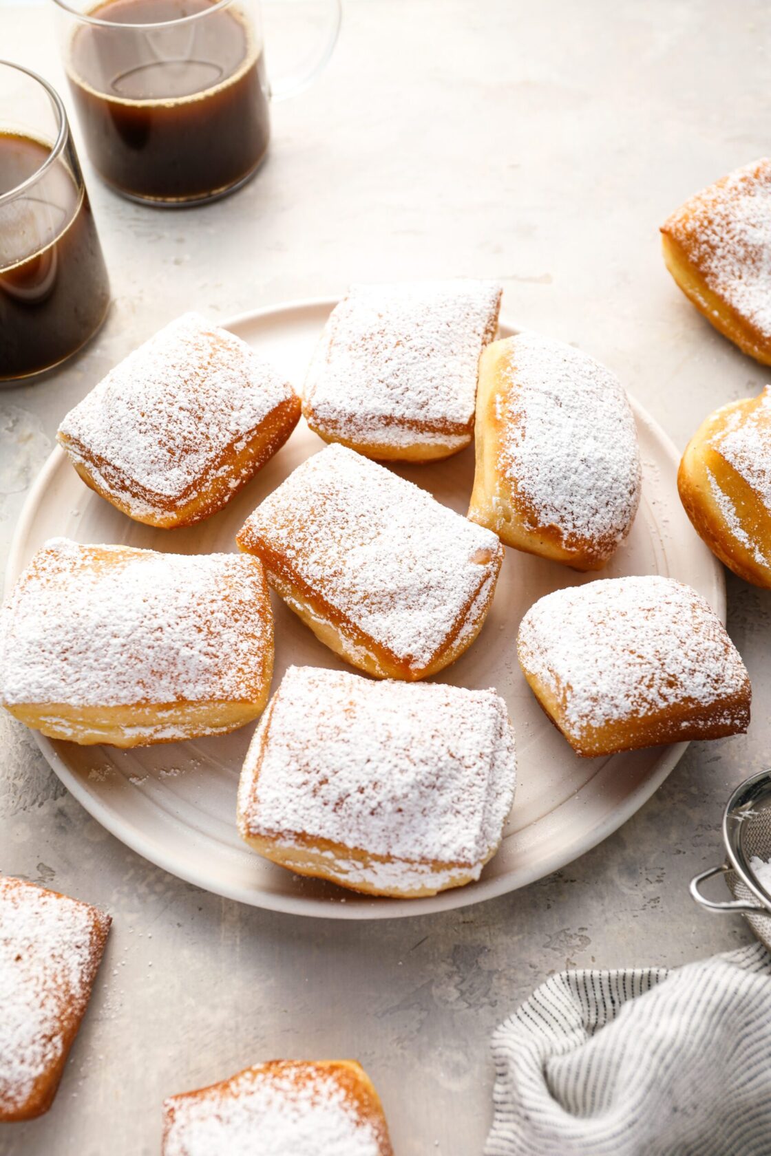 sugar-dusted beignets on round white plate with 2 cups of coffee