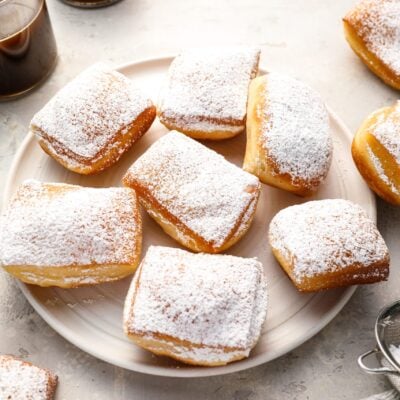 sugar-dusted beignets on round white plate with 2 cups of coffee