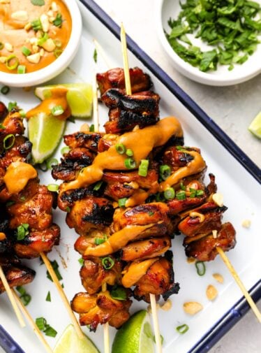 white serving platter with cooked chicken skewers and small bowl of peanut sauce
