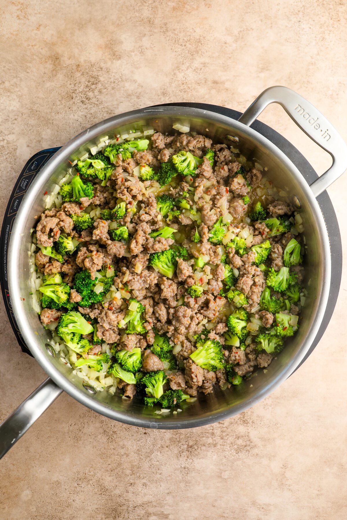 large skillet with onion, garlic, broccoli florets, and raw sausage