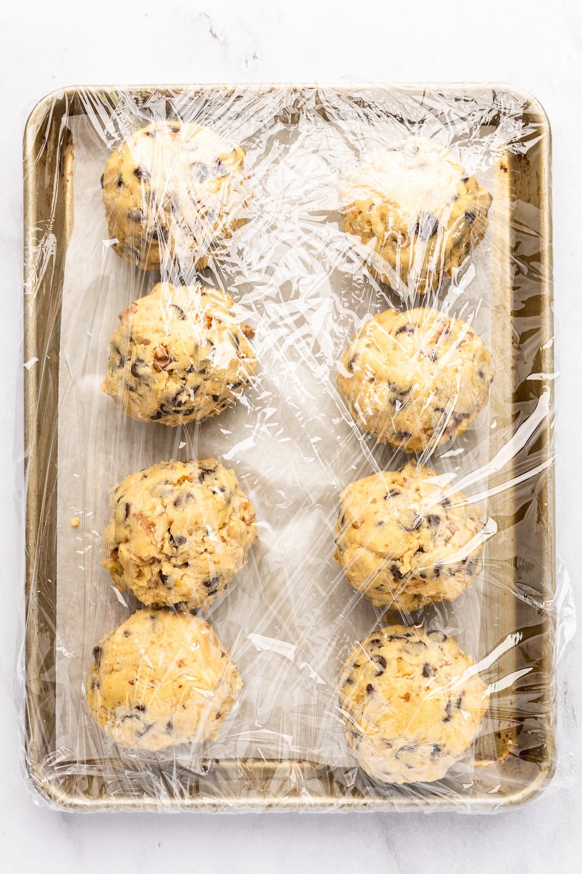 parchment-lined baking sheet with cookie dough divided into 8 balls and covered with plastic wrap