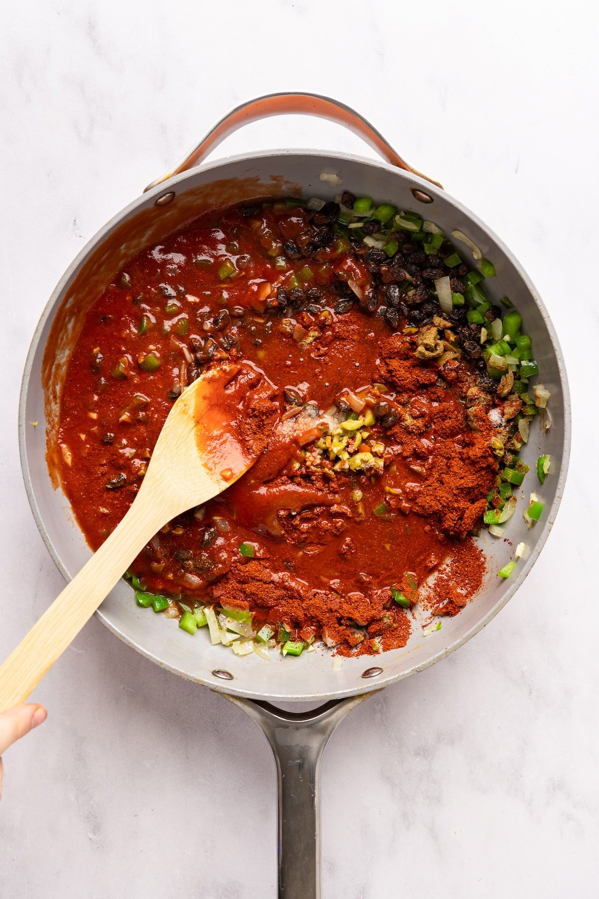 large skillet with tomato sauce, broth, raisins, sazon seasoning, salt, cumin, brown sugar, and olives added to onion/pepper/garlic mixture