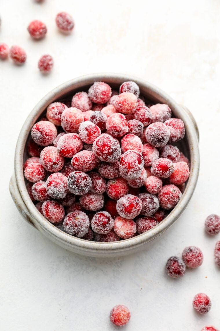 sugared cranberries in white bowl with scattered cranberries surounding bowl