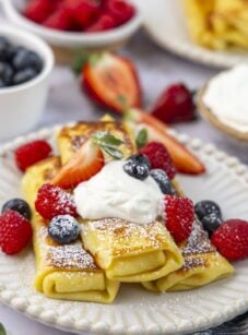 white plate with 3 cheese blintzes topped with sour cream, confectioners' sugar, and fresh berries