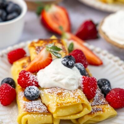 white plate with 3 cheese blintzes topped with sour cream, confectioners' sugar, and fresh berries