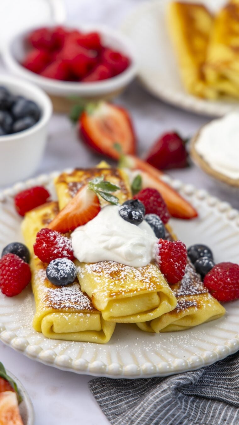 white plate with 3 cheese blintzes topped with sour cream, confectioners' sugar, and fresh berries