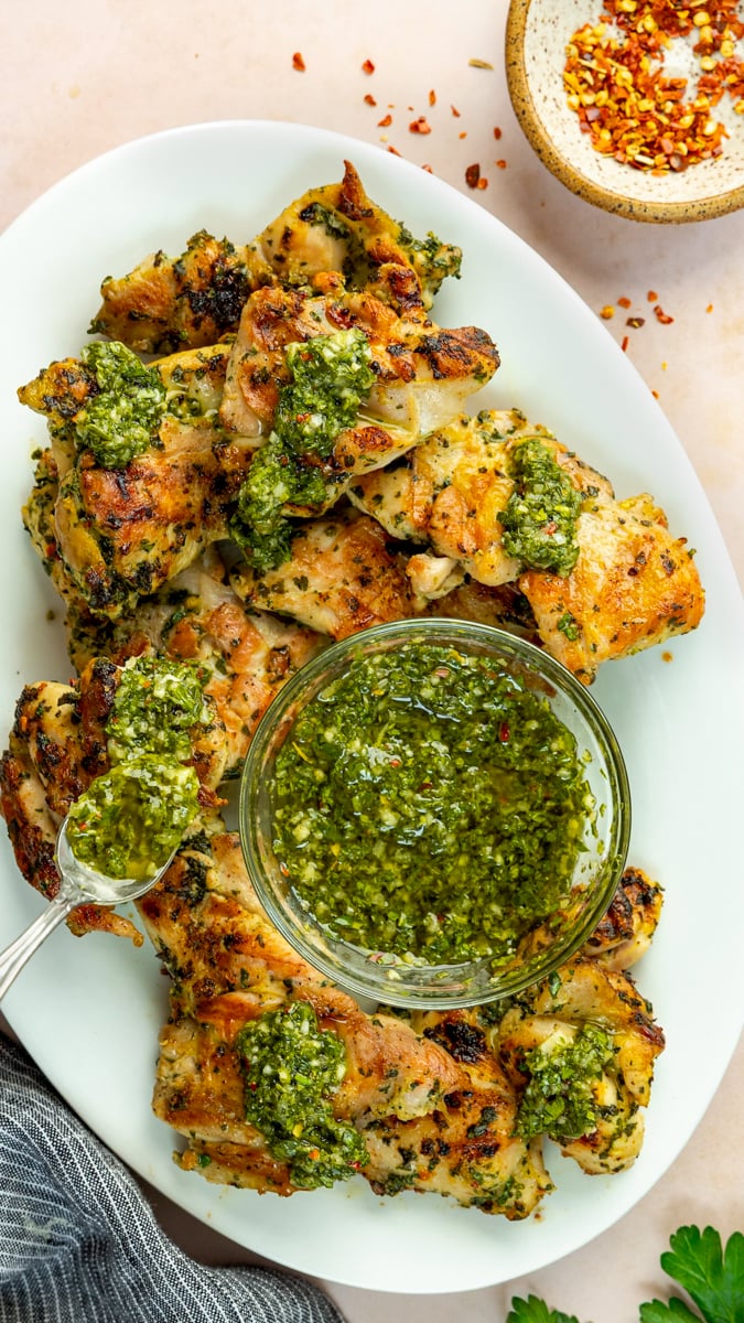 Grilled chimichurri chicken thighs on platter with chimichurri sauce.