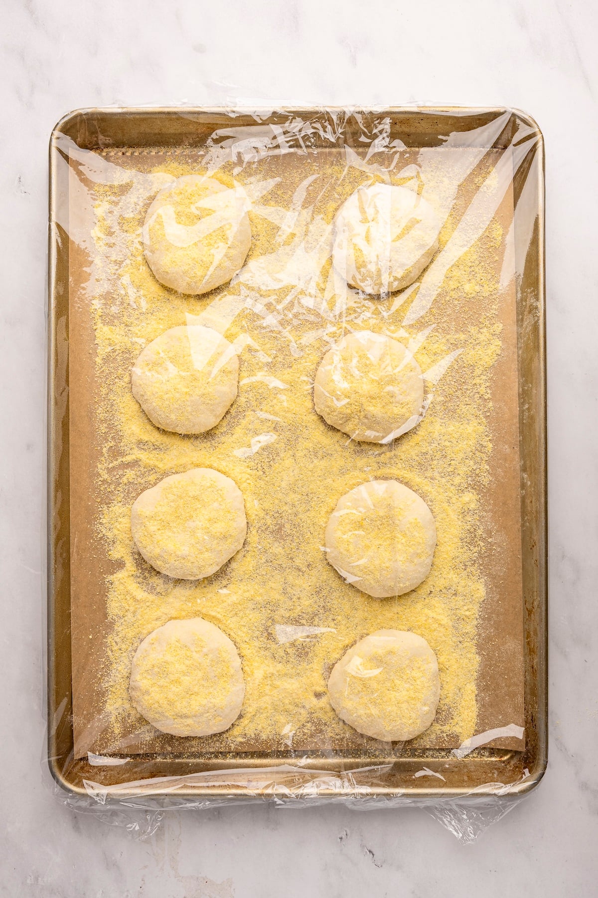 Parchment-lined baking sheet with plastic wrap over English muffin rounds